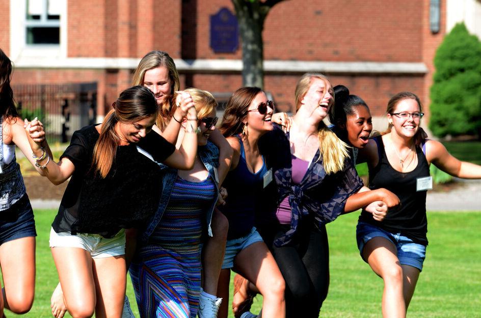 A group of female students hold hands while participating in an orientation activity on the campus lawn